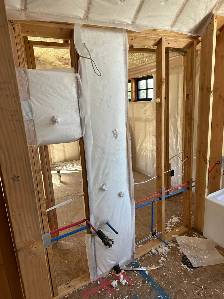 Fiberglass batt insulation being installed around PEX plumbing lines, vapor barrier visible, interior walls being insulated