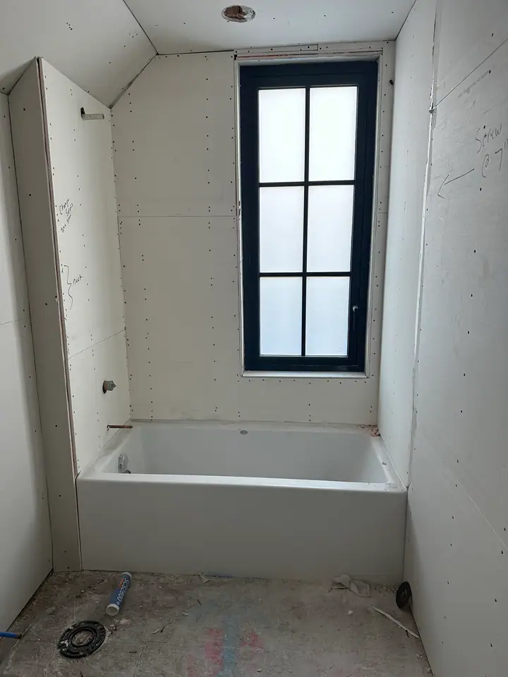 Bathroom taking shape, tub set in place, drywall hung, black-frame window installed, plumbing complete