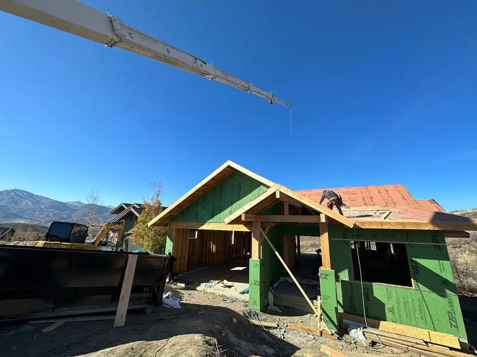 Wide shot of house framing with roof trusses, crane arm visible, and mountains in the background