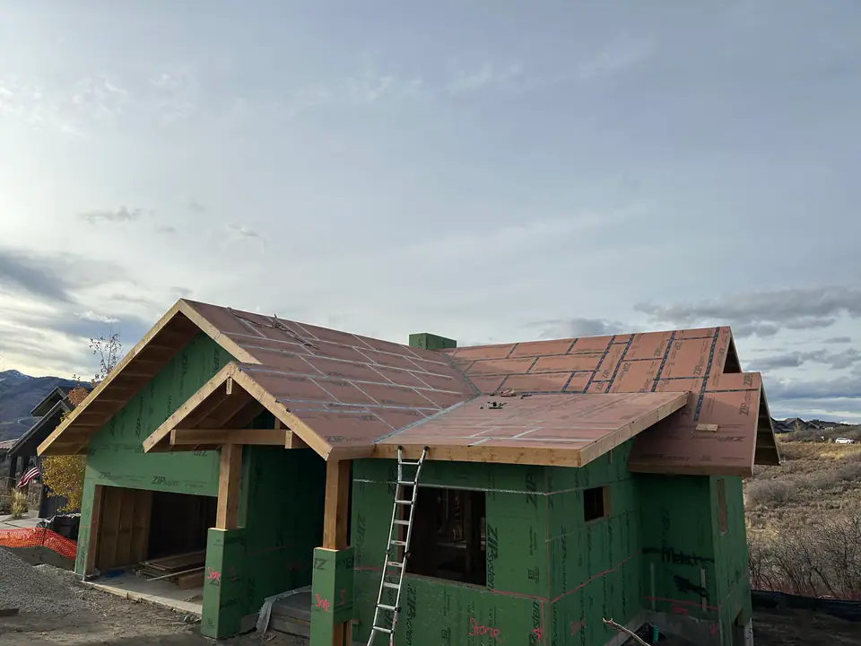 Full exterior view of the home during framing, ZIP System green sheathing on walls, ZIP-R roof panels, timber gable entry, ladder against the side, fall colors on the hillside