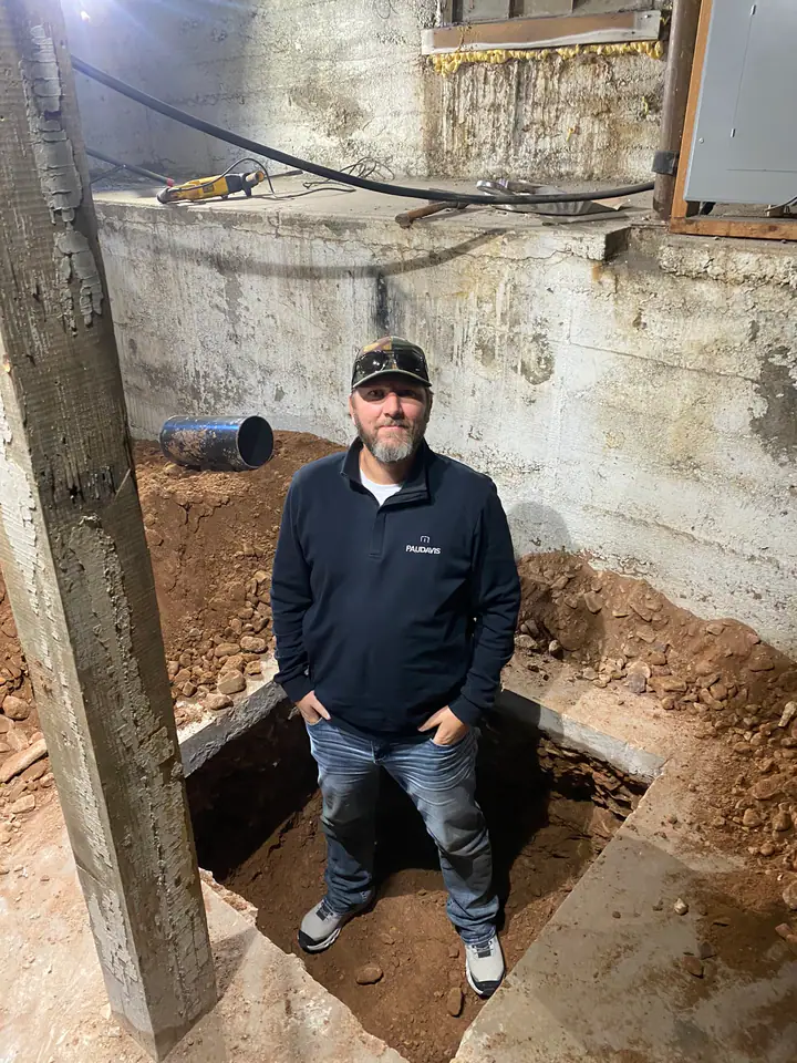 Nate Jensen standing in a deep footing excavation pit next to the foundation wall, demonstrating the depth of underpinning work required for structural repair