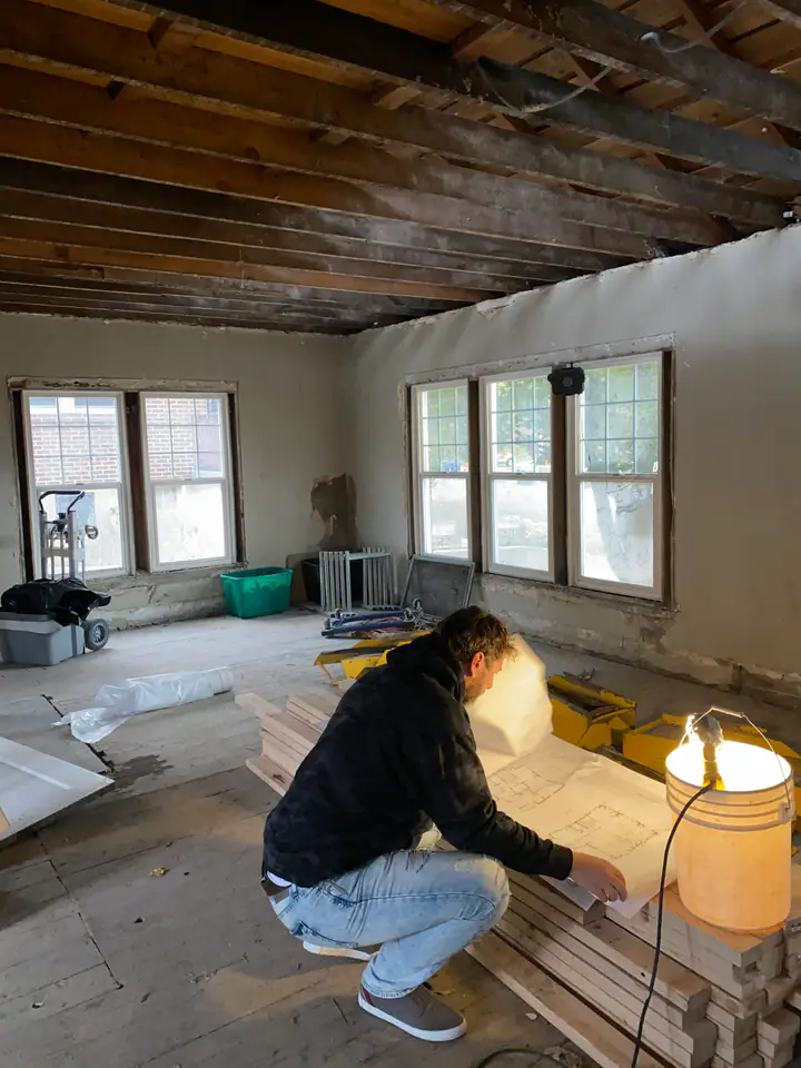 Nate Jensen reviewing structural blueprints inside a gutted older home, exposed ceiling joists and natural light through original windows