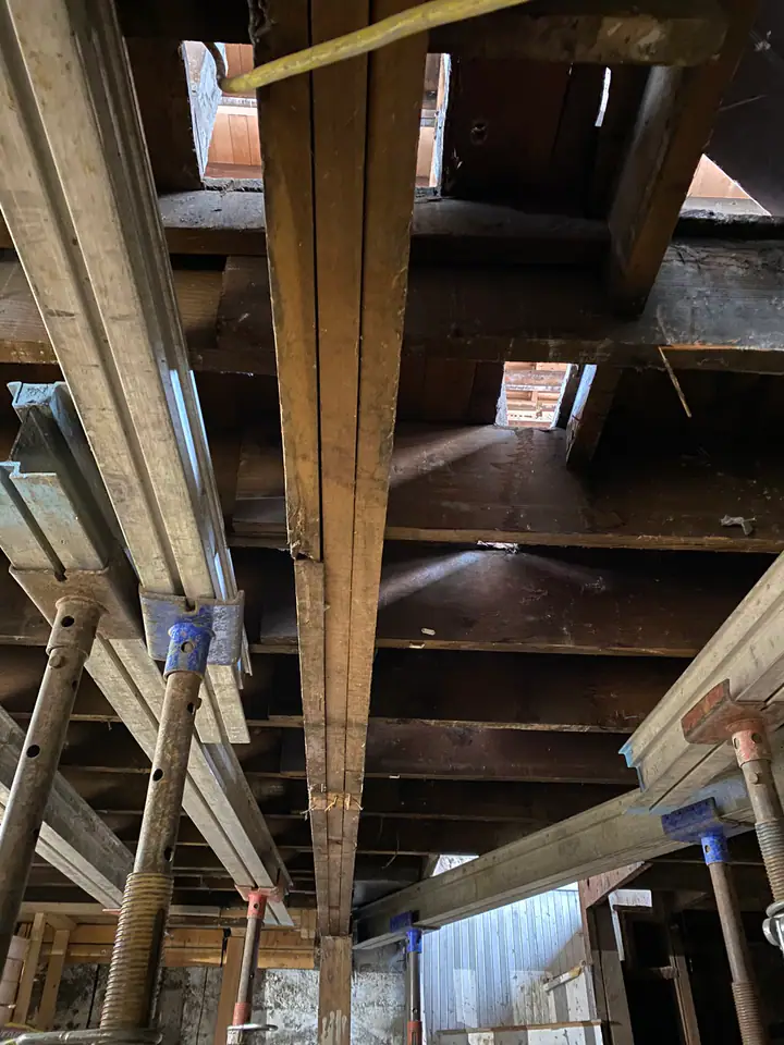 Looking up through floor joists at temporary steel shoring system, gaps in subfloor visible showing the extent of structural compromise