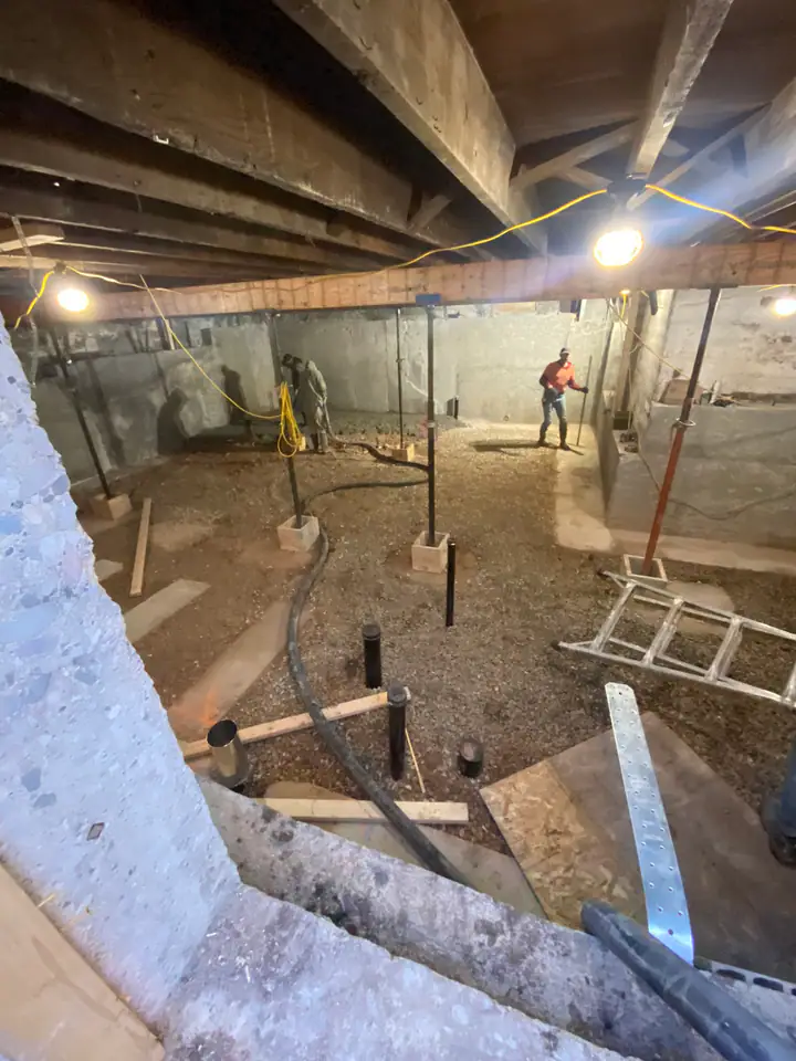 Wide view of crawlspace being rehabilitated, new concrete walls poured, steel posts on pad footings, worker in orange hi-vis