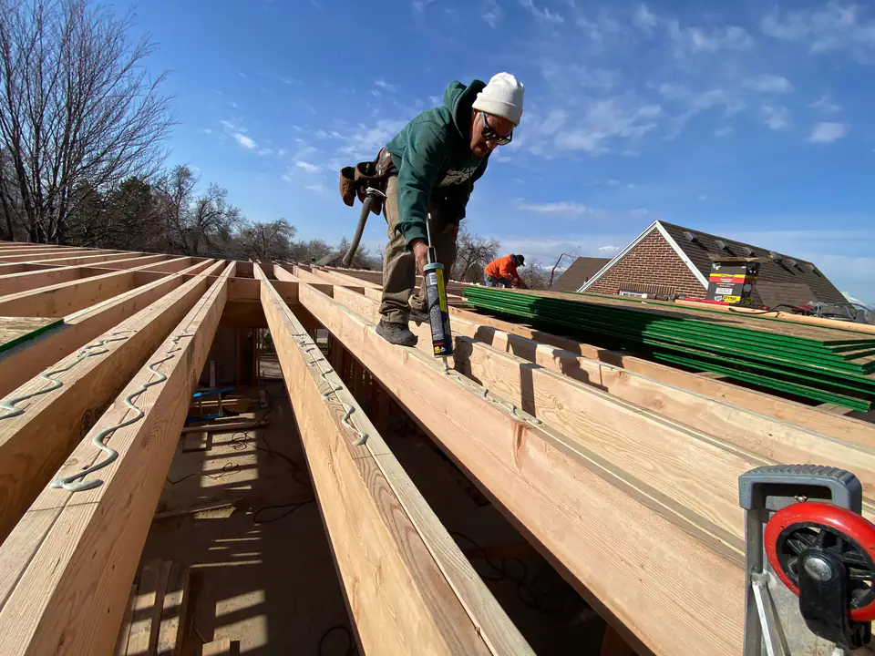 Worker with caulk gun sealing new floor joists, mountains visible in background, precision craftsmanship in every connection