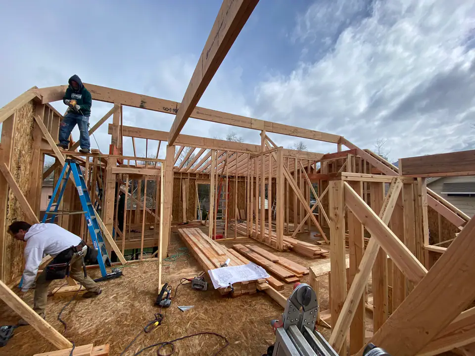 Wide angle of upper level framing, workers on ladders, ridge beam visible, blueprints on floor, dramatic clouds through open walls