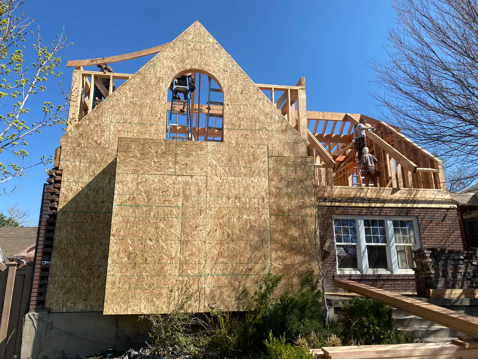 Full exterior view of new gable end wall with arched window opening, OSB sheathing complete, workers on ladders, massive scale of addition visible next to original brick