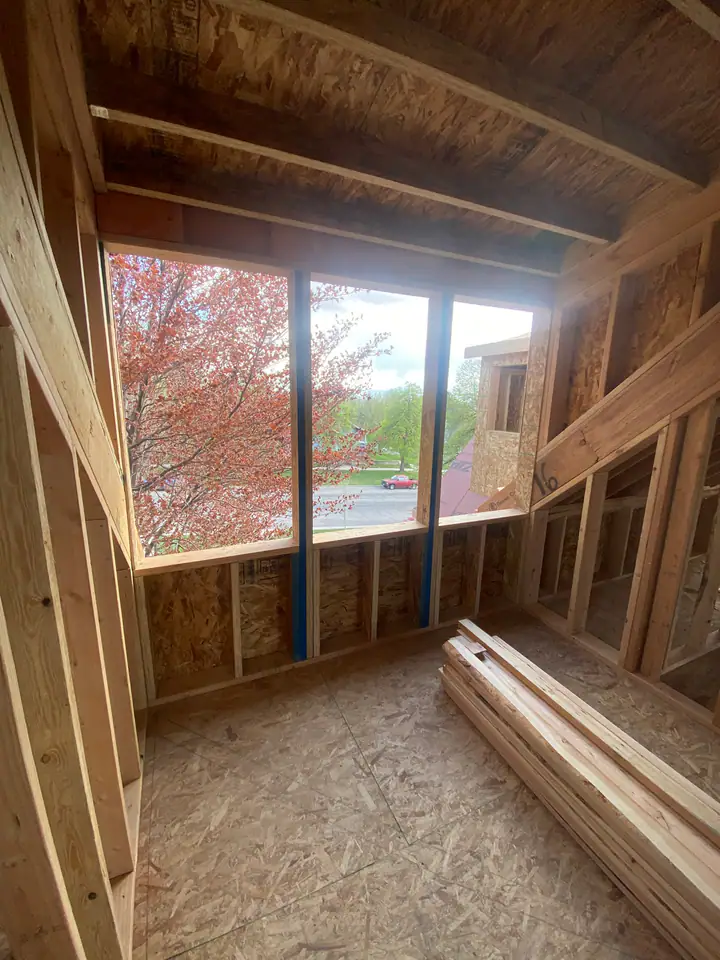 New upper level room with large window openings framed, OSB floor, vaulted ceiling, flowering tree visible outside